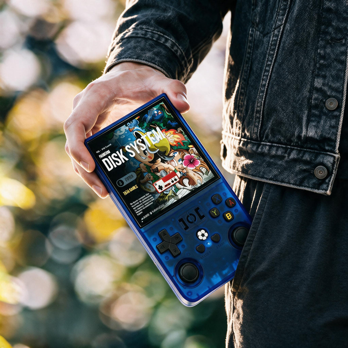 A person in a dark denim jacket holds r36max translucent blue retro handheld gaming console with a colorful screen displaying 'DISK SYSTEM'