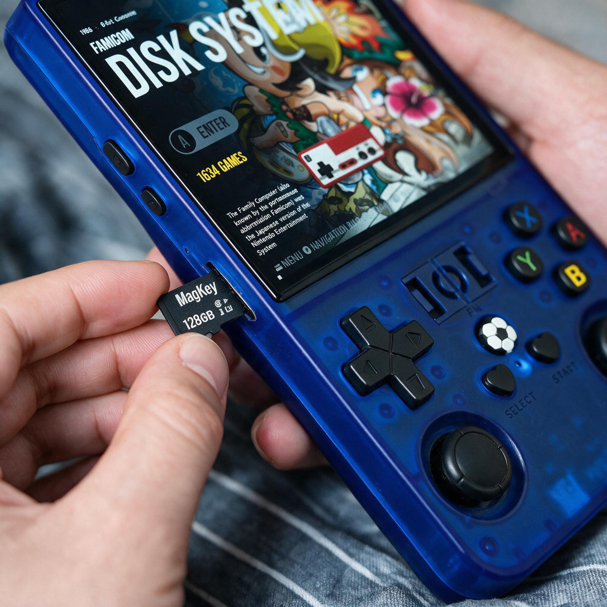 A close-up of a r36max blue handheld gaming console being held in two hands, with a small black MagSafe-compatible 2285 MB microSD card being inserted near the screen. The console screen displays a colorful 'Famicom Disk System' game menu.
